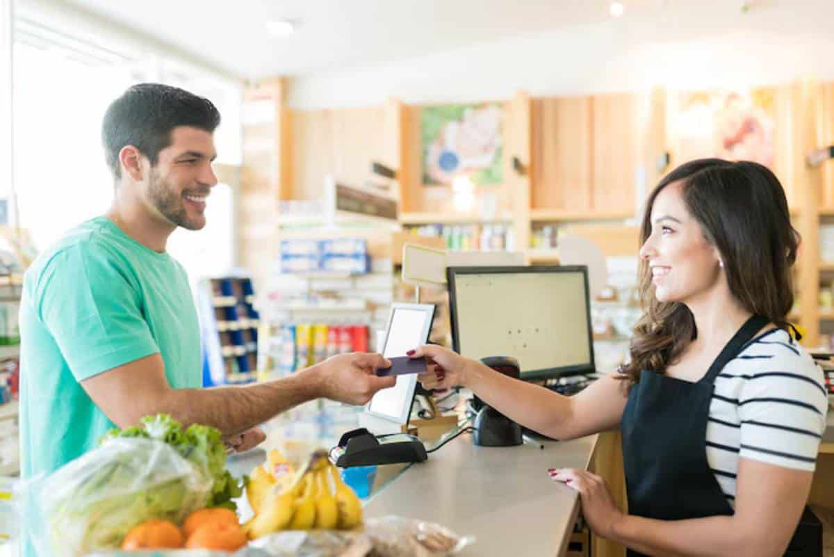 A customer in a green shirt hands a card to a cashier at a grocery store, with fruits and vegetables on the counter.