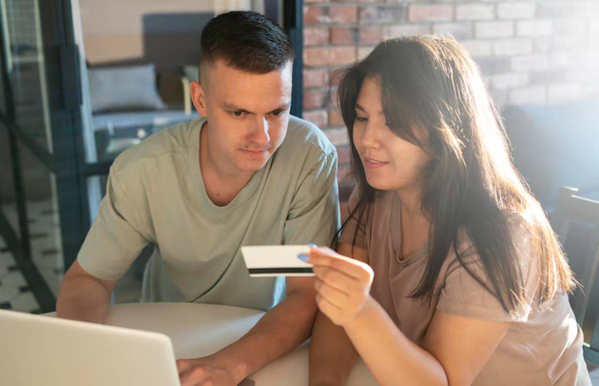 A woman holds a credit card while sitting beside a man, both focused on a laptop in a brightly lit, modern interior space.