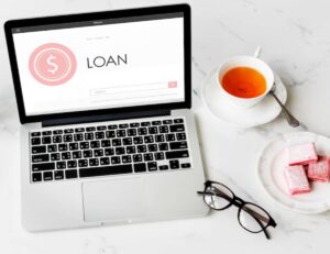 A laptop displaying a loan search page, beside a cup of tea, plate of pink sweets, and a pair of glasses on a marble surface.