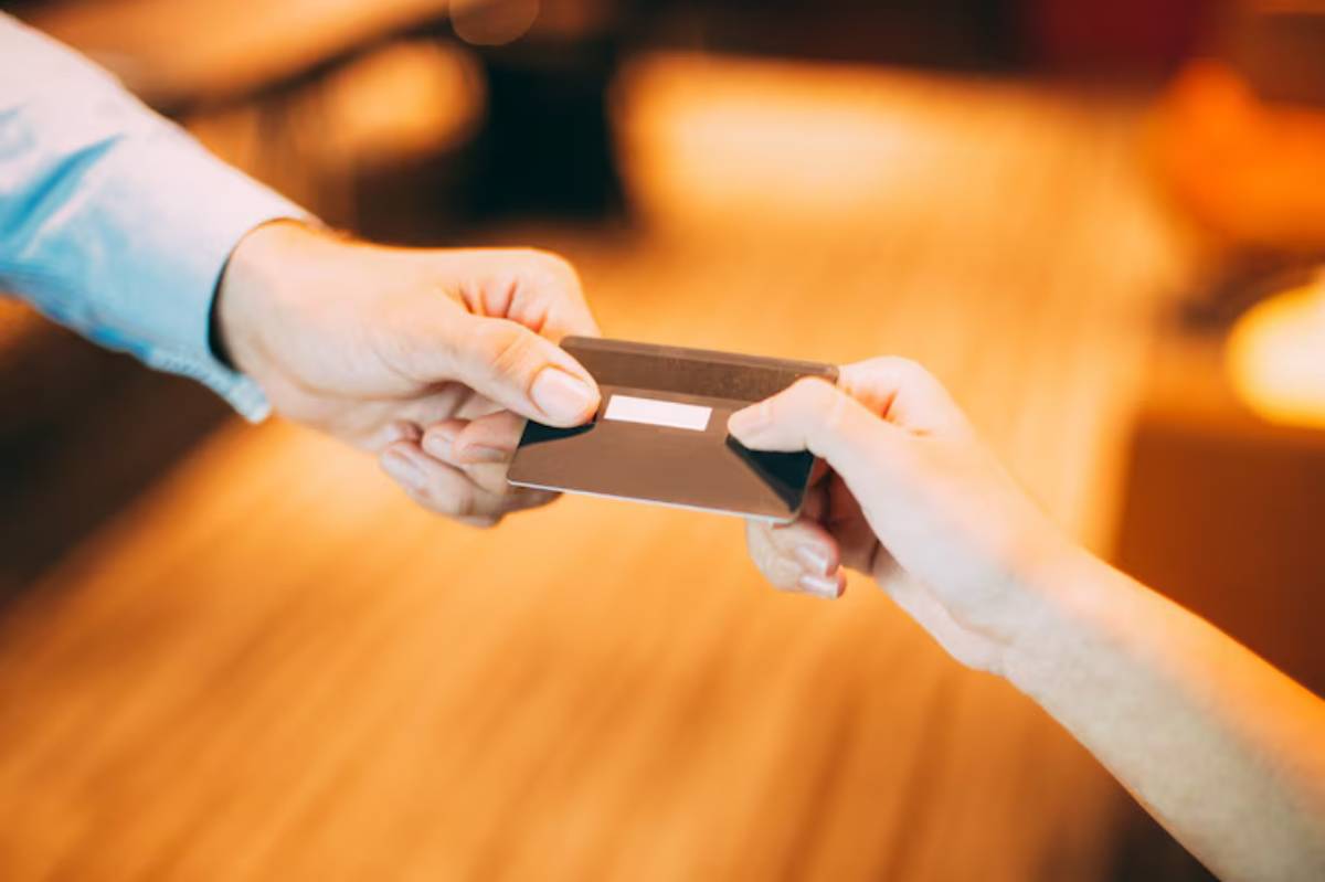 A hand transfers a black credit card to another hand, symbolizing a financial exchange in a warm, blurred indoor setting.