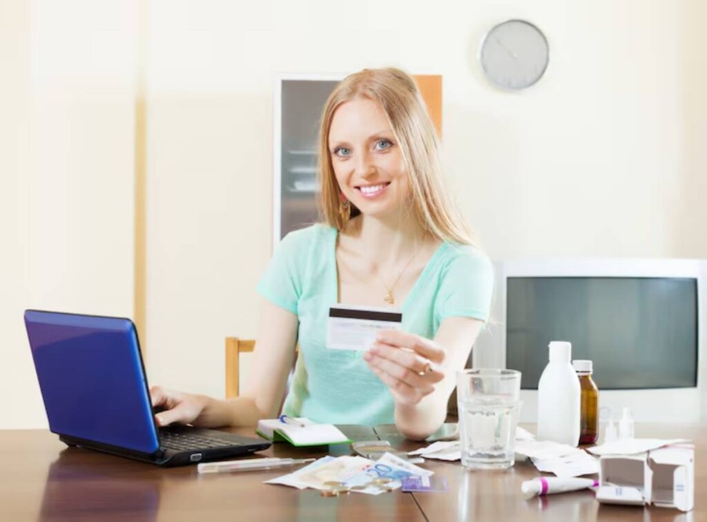 A woman sits at a table with a laptop, holding a credit card, surrounded by papers, medicine bottles, and a glass of water.
