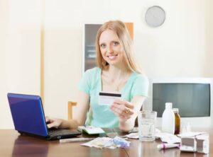 A woman sits at a table with a laptop, holding a credit card, surrounded by papers, medicine bottles, and a glass of water.