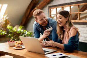 A couple interacts at a wooden table, working on a laptop, surrounded by snacks and plants in a cozy room.