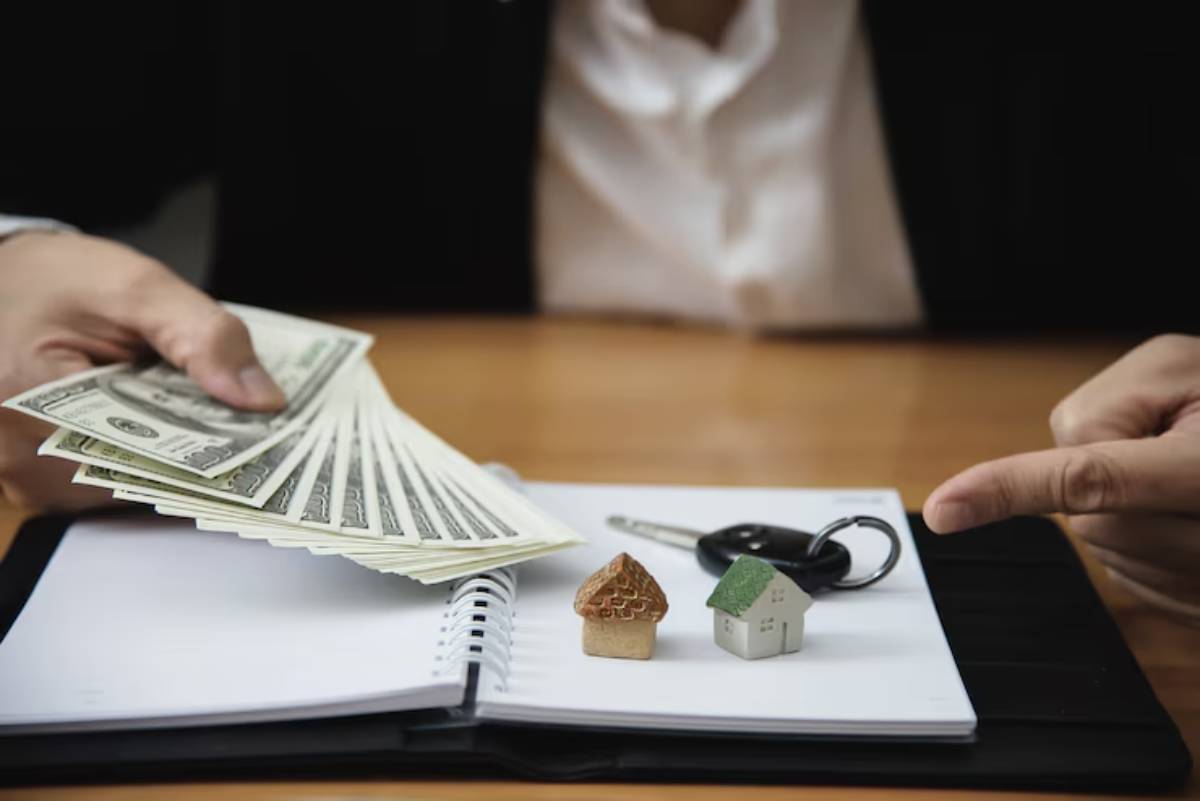 A hand offers cash above a notebook, with two small house models and car keys, symbolizing real estate and transactions.
