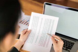 A person analyzing financial data on a printed spreadsheet while holding a pen, with a laptop and charts in the background.