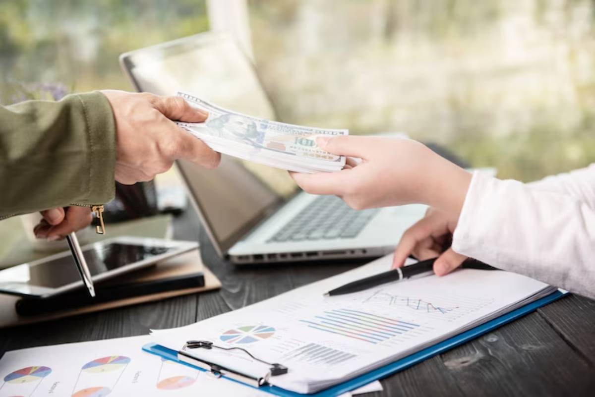 A person hands cash to another over a desk cluttered with financial documents and a laptop. 