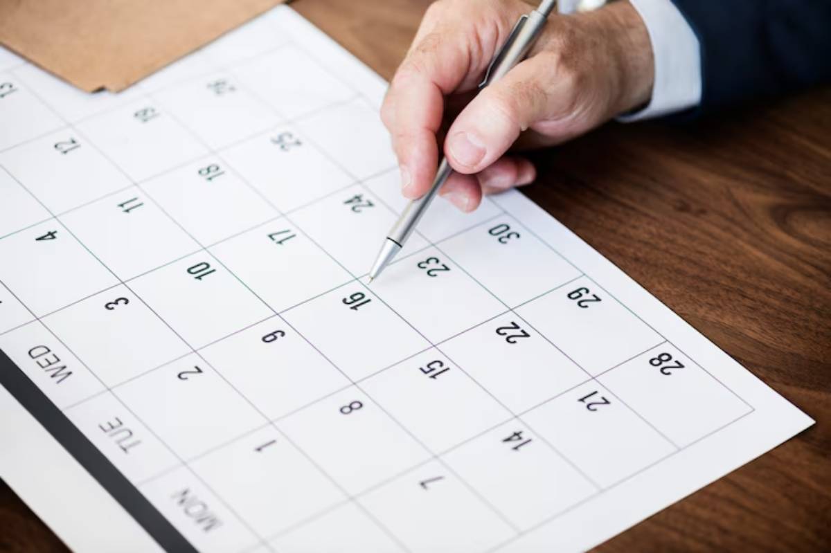A hand holding a pen marks a date on a printed calendar laid on a wooden table.