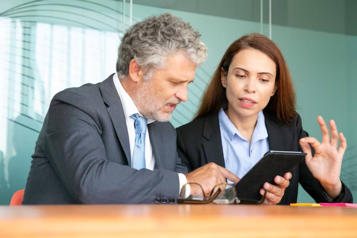 Two business professionals engaged in discussion over a tablet, seated at a conference table in a modern office setting.