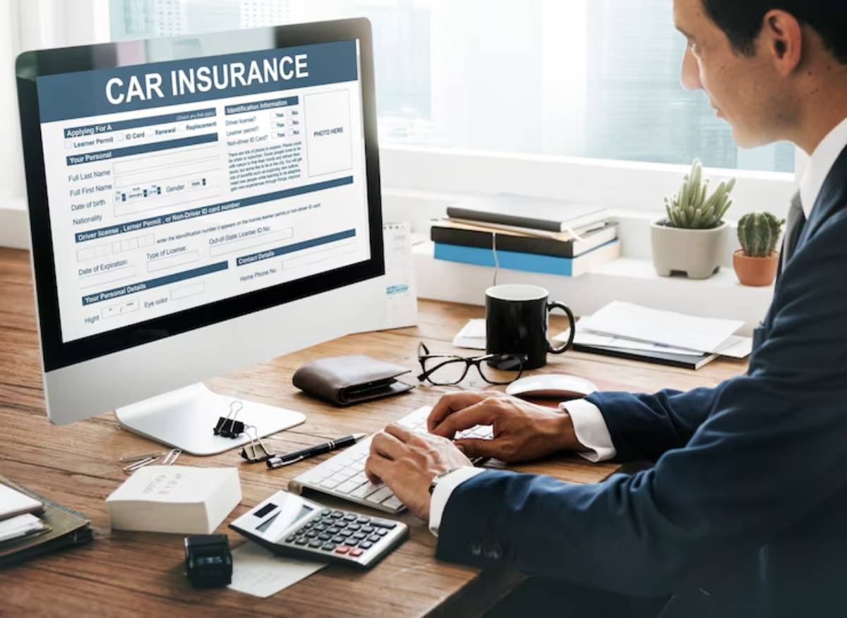 A person in a suit is typing at a desk with a computer displaying a car insurance form, surrounded by office supplies and plants.