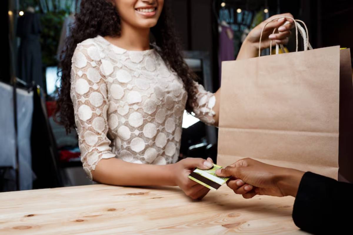 A smiling woman in a white blouse hands over a credit card while receiving a paper shopping bag at a checkout counter. 