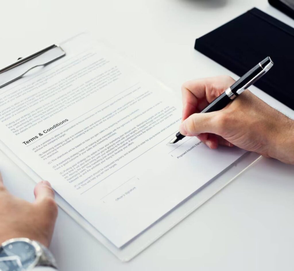 A hand holding a fountain pen signs a document titled "Terms & Conditions" on a clipboard with a watch visible on the wrist.