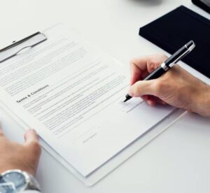 A hand holding a fountain pen signs a document titled "Terms & Conditions" on a clipboard with a watch visible on the wrist.