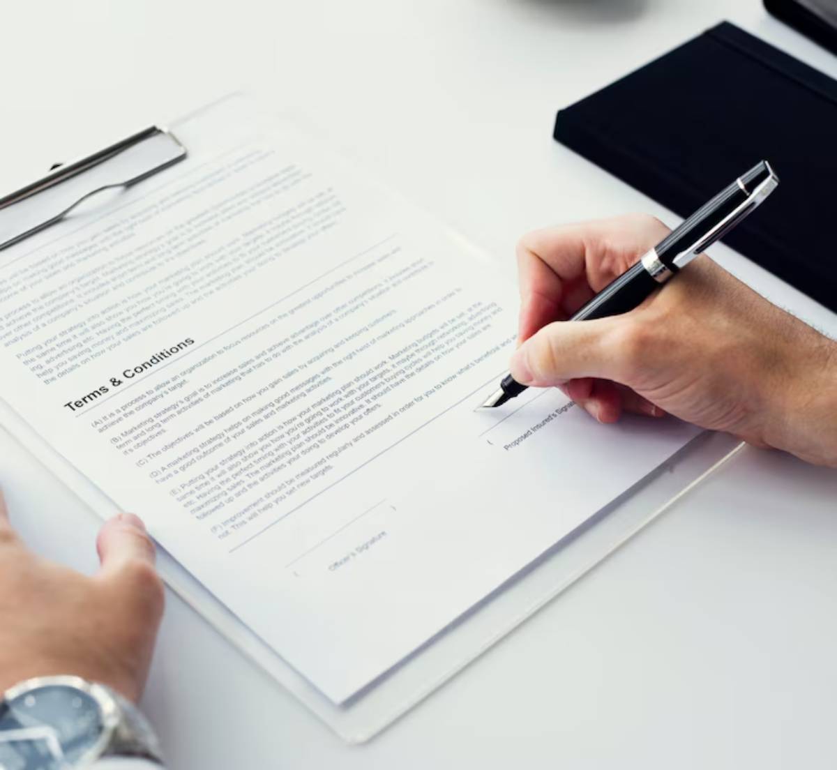 A hand holding a fountain pen signs a document titled "Terms & Conditions" on a clipboard with a watch visible on the wrist.