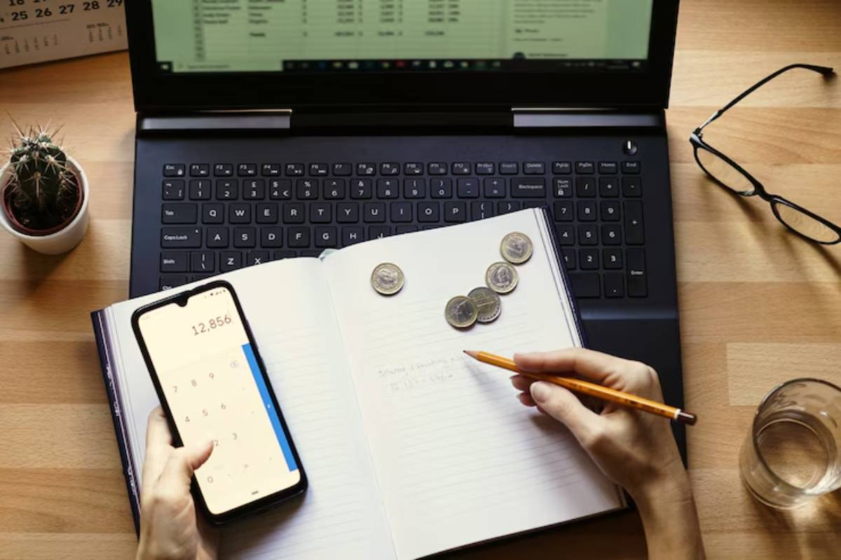 A person calculates finances using a smartphone calculator, writing in a notebook, with coins and a laptop in the background. 