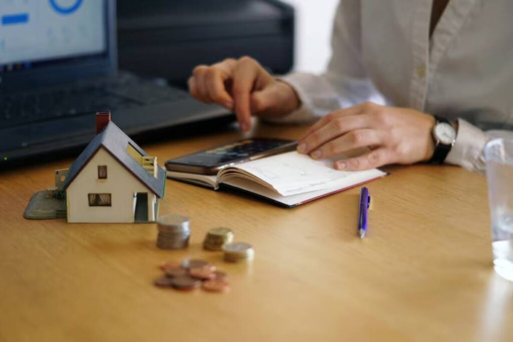 A person calculates finances on a smartphone, surrounded by a model house, coins, notes, and a laptop in a home office setting.