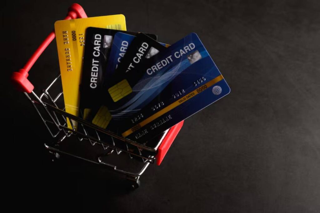 A small shopping cart holds multiple credit cards, including a yellow one, against a dark background.