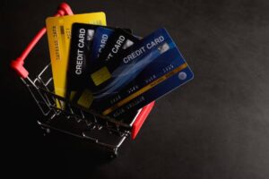 A small shopping cart holds multiple credit cards, including a yellow one, against a dark background.