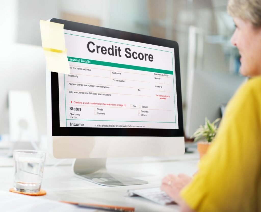 A person sits at a desk typing on a computer displaying a Credit Score form with fields for personal details and income status.