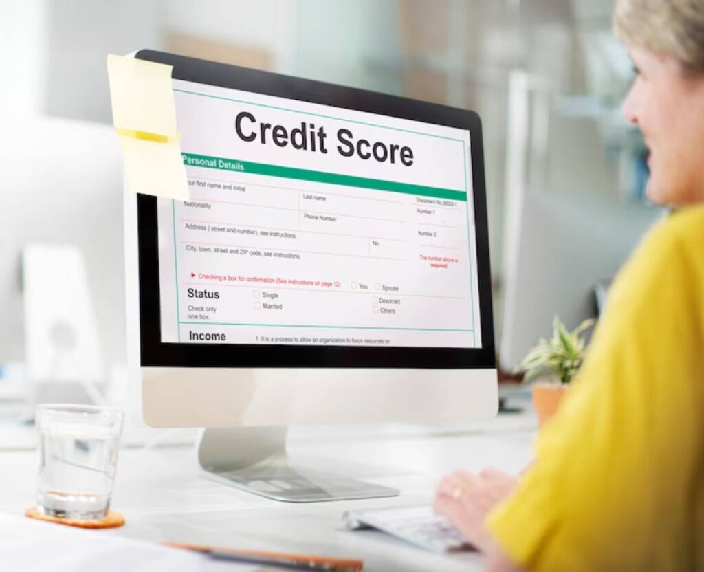 A desktop computer displays a Credit Score form with sections for personal details, status, and income, beside a glass of water.