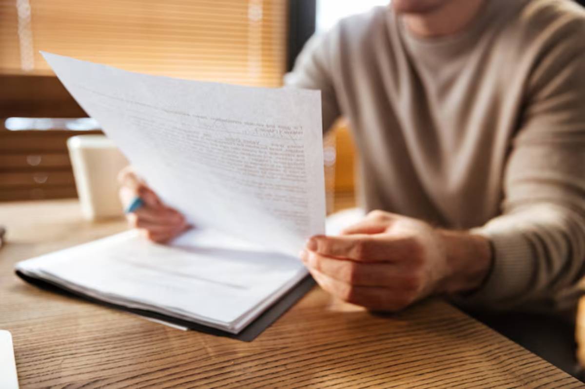 A person in a light sweater holds a page of documents, examining it closely at a wooden table in a well-lit setting. 