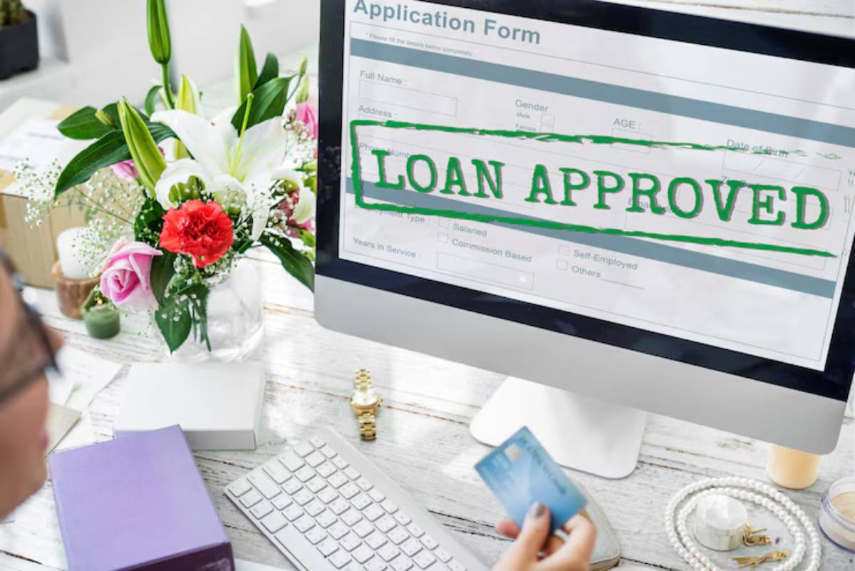 A person holds a credit card, looking at a computer screen displaying an LOAN APPROVED message above an application form. Floral arrangement nearby. 