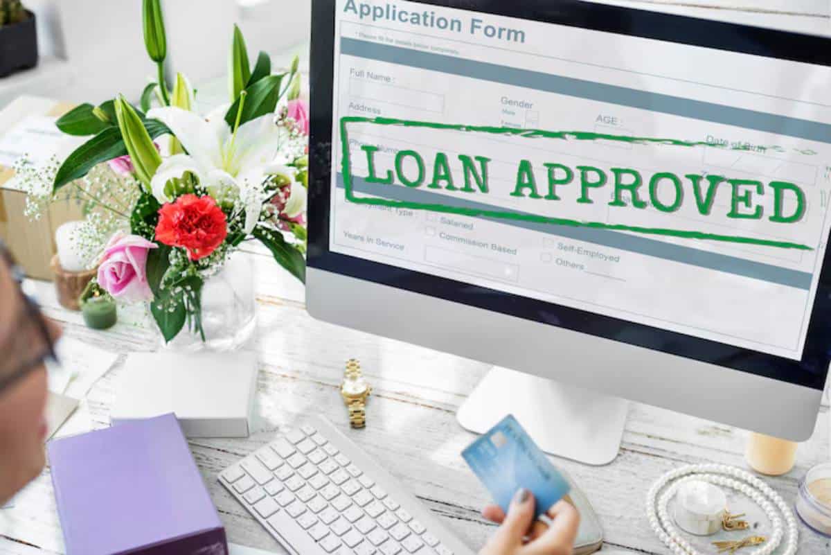 A person holds a credit card, looking at a computer screen displaying an LOAN APPROVED message above an application form. Floral arrangement nearby.