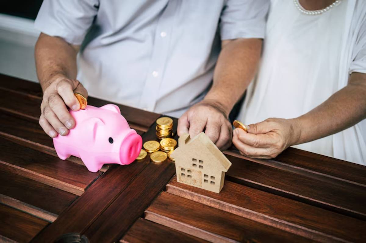 A couple saves money by adding coins to a pink piggy bank, next to a model house and stacks of gold coins on a wooden table.