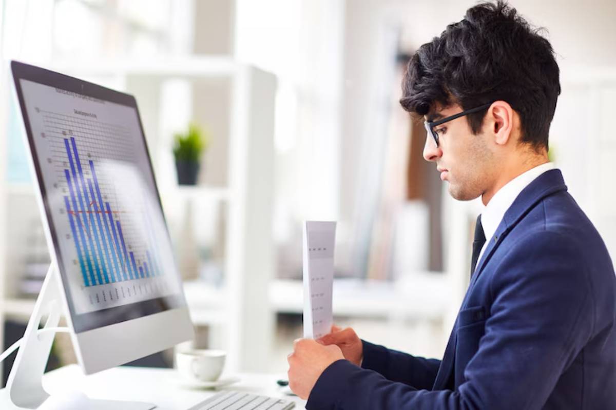 A professional man in a suit sits at a desk, reviewing a printed report while analyzing graphs on a computer screen. 