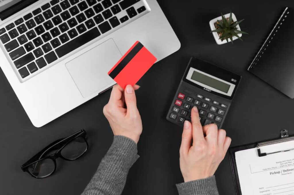 A person's hand holds a red credit card while calculating expenses on a black calculator, with a laptop and paperwork nearby.