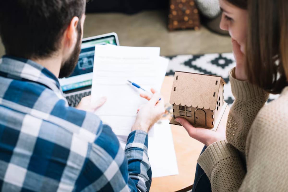 A person seated at a table reviews documents while another holds a small wooden house model, showcasing a planning or creative session. 