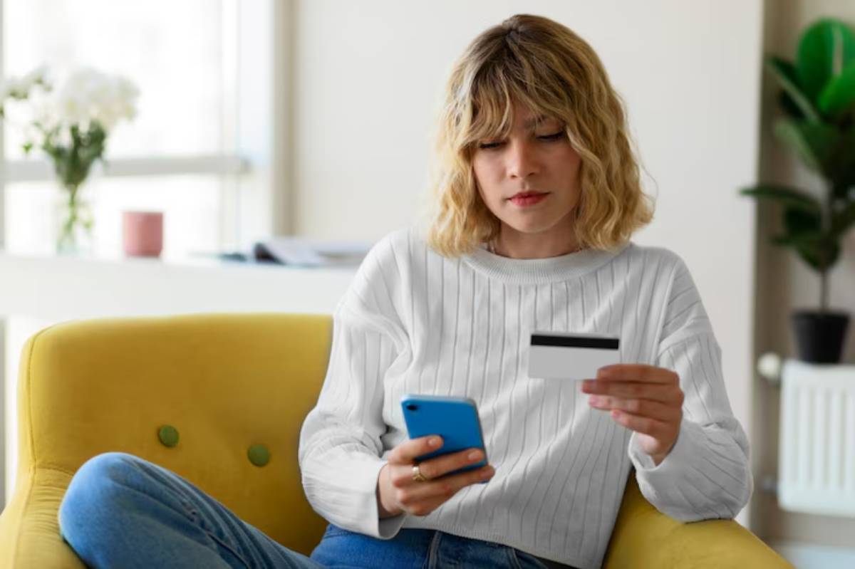 A person sits on a yellow couch, holding a smartphone in one hand and a credit card in the other, surrounded by a bright interior. 