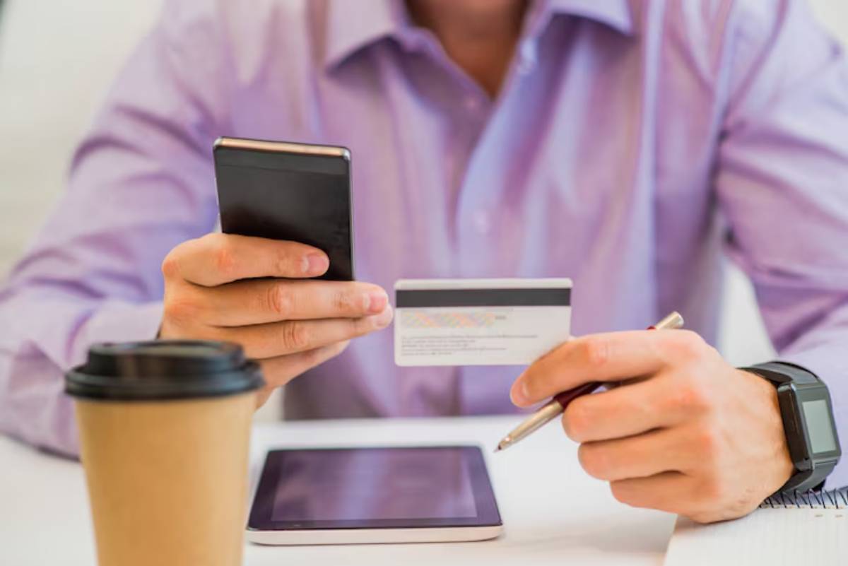 A person in a purple shirt holds a smartphone and credit card, ready to make an online payment, with a coffee cup nearby. 