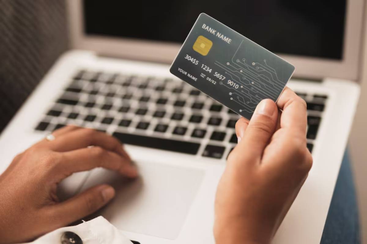 A person holding a bank card in one hand while using a laptop, indicating online shopping or banking activity.