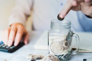 A person drops a coin into a glass jar filled with banknotes, while using a calculator on a desk.