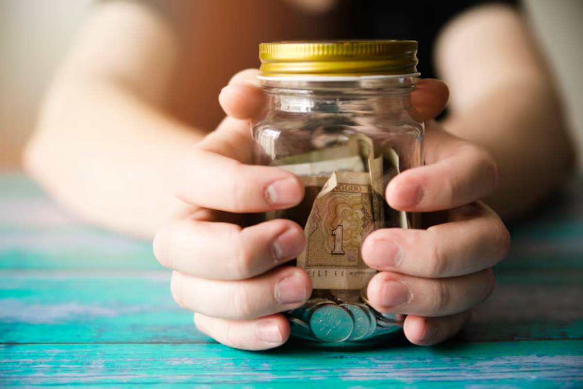 A person's hands gently hold a jar filled with coins and folded bills on a turquoise wooden surface, symbolizing savings. 