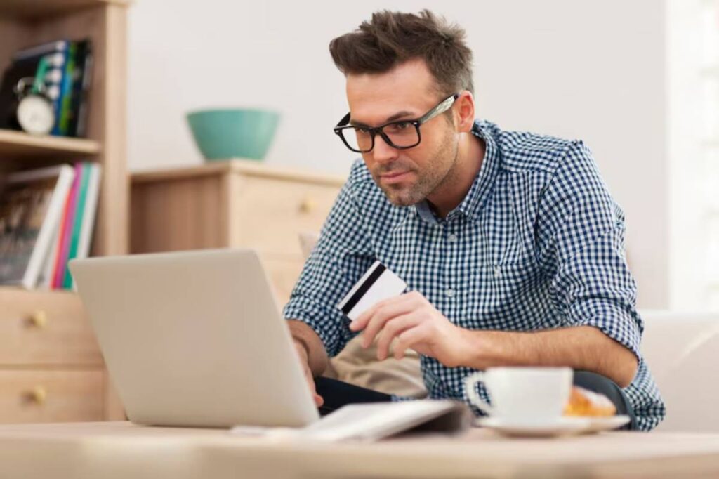 A person sits on a couch, focused on a laptop, holding a credit card in their other hand, with a cup and pastry nearby.