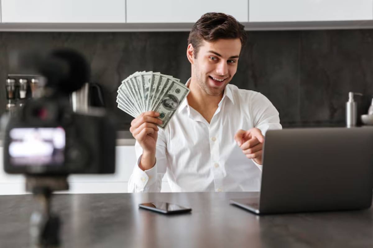 A person in a white shirt holds a fan of cash while pointing, sitting in front of a laptop and camera on a kitchen table.