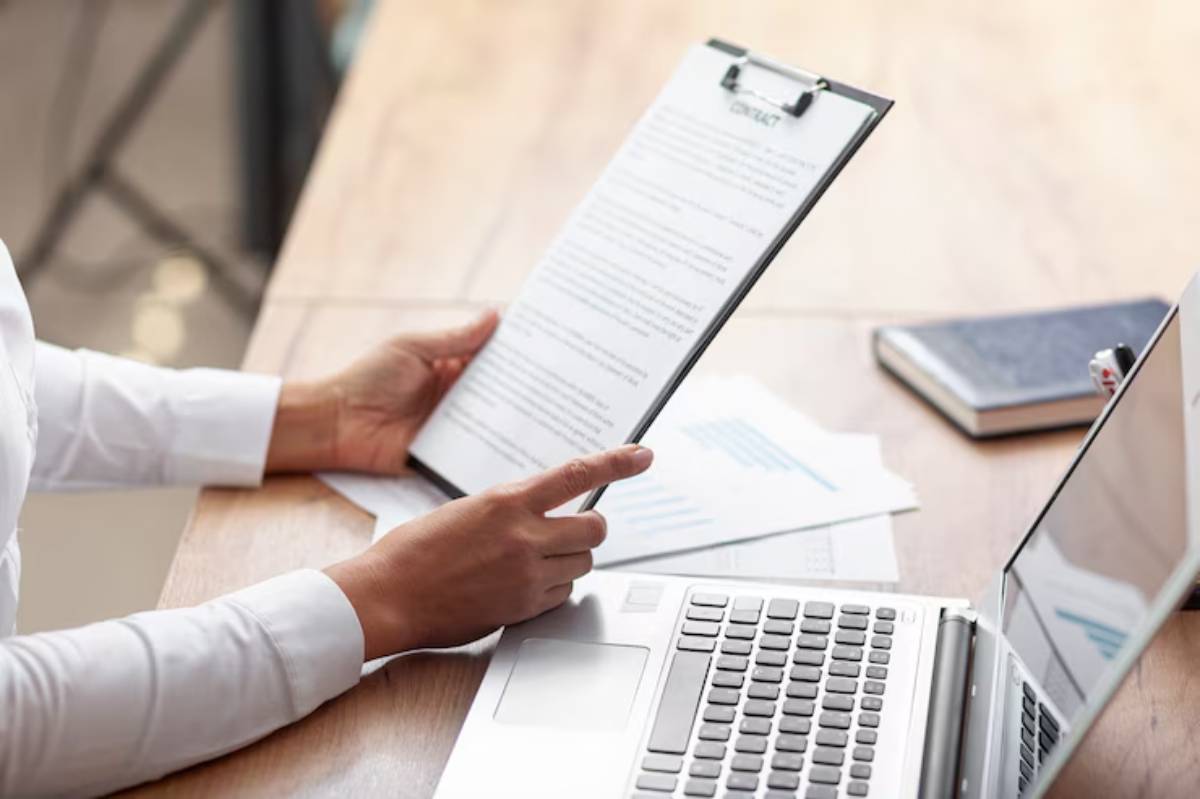 A person holding a clipboard with a document, sitting at a wooden desk with a laptop and scattered papers. 