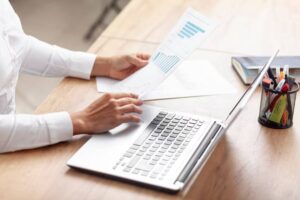 A person reviewing printed charts and graphs while working on a laptop at a wooden desk, surrounded by stationery and a notebook.