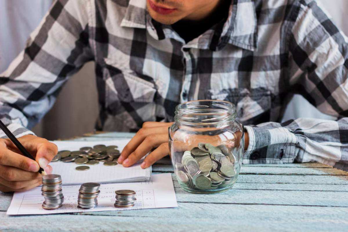  A person in a plaid shirt counts coins and takes notes, with stacked coins and a jar of coins on a wooden table.