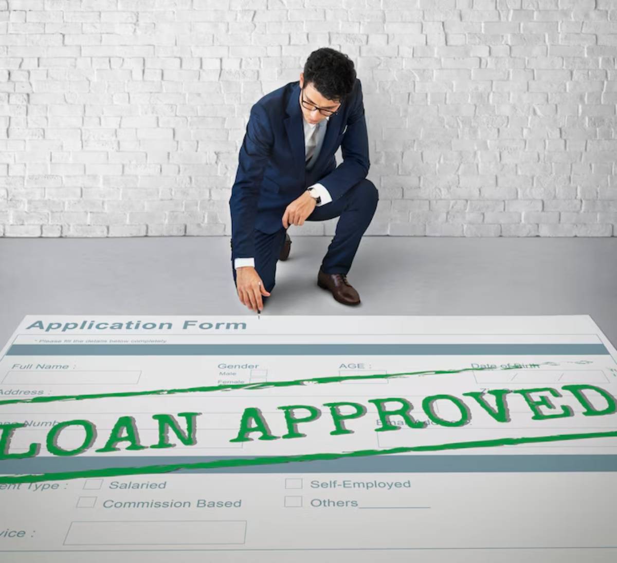 A man in a suit kneels beside a large application form marked with LOAN APPROVED in bold green letters on a gray floor. 