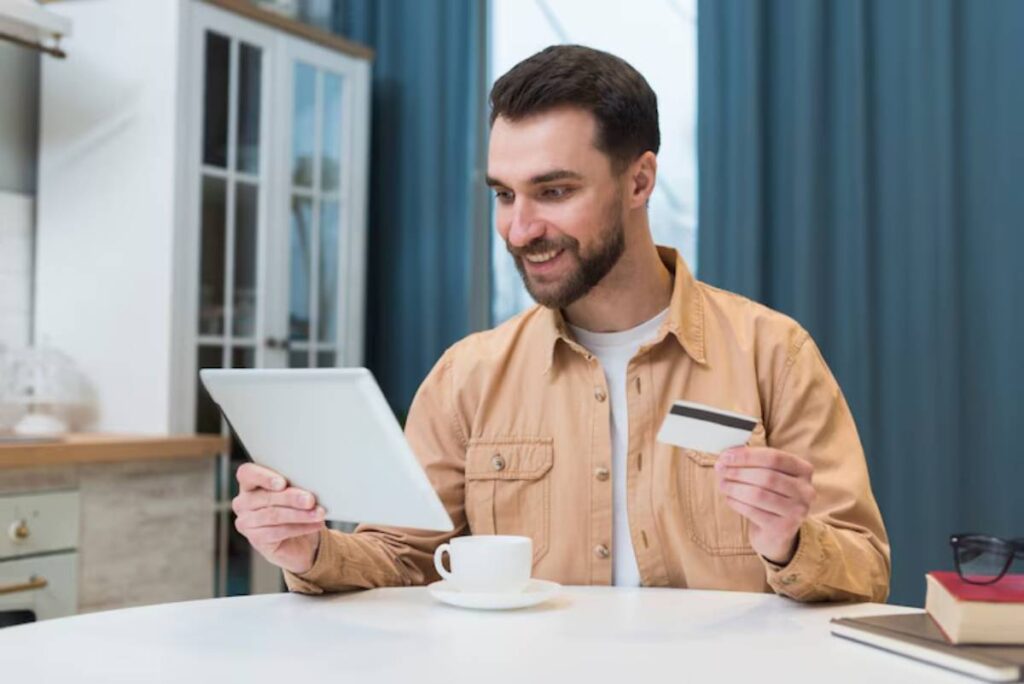 A person in a beige shirt is seated at a table, holding a tablet in one hand and a credit card in the other, with a coffee cup nearby.
