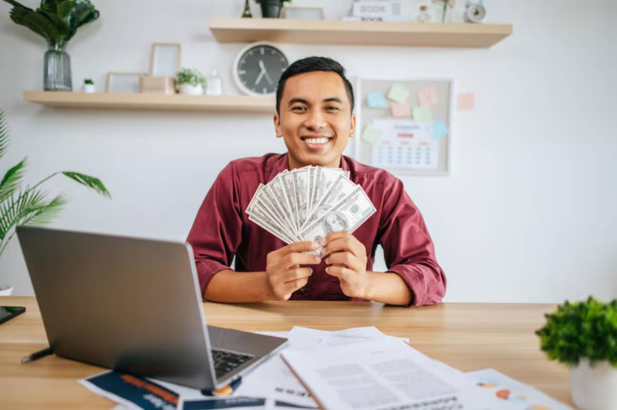 A person in a red shirt holds a fan of money in front of a laptop and documents on a desk, with plants in the background.
