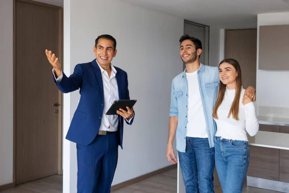 A real estate agent presents a modern apartment to a couple, showcasing an open living area with neutral colors and wooden flooring.