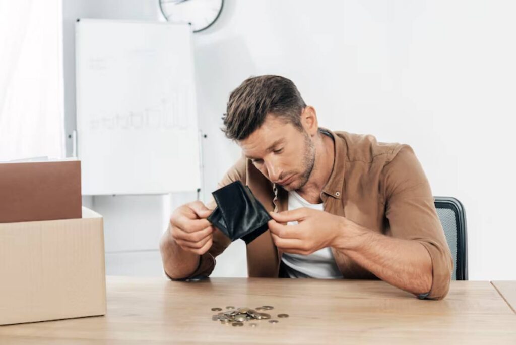 A man examines an empty wallet, surrounded by coins and a cardboard box, in a bright, minimalistic office space.
