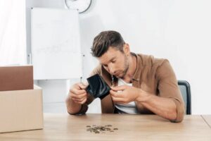 A man examines an empty wallet, surrounded by coins and a cardboard box, in a bright, minimalistic office space.