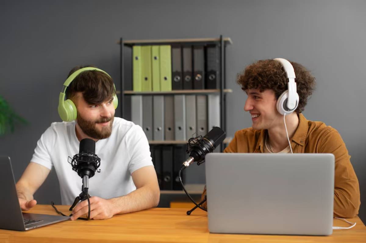  Two individuals wearing headphones sit at a wooden desk with microphones and laptops, surrounded by office binders in a modern workspace.