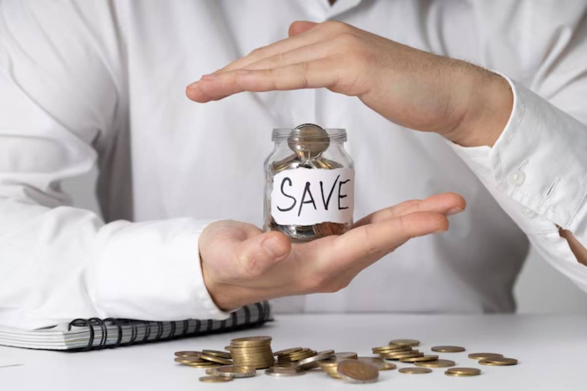 A person in a white shirt holds a jar labeled SAVE with coins inside, surrounded by scattered coins on a table. 