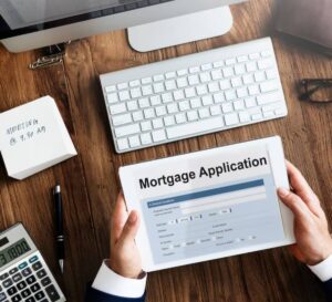 A person in a suit holds a tablet displaying a mortgage application form, with a computer and notepad on a wooden desk.