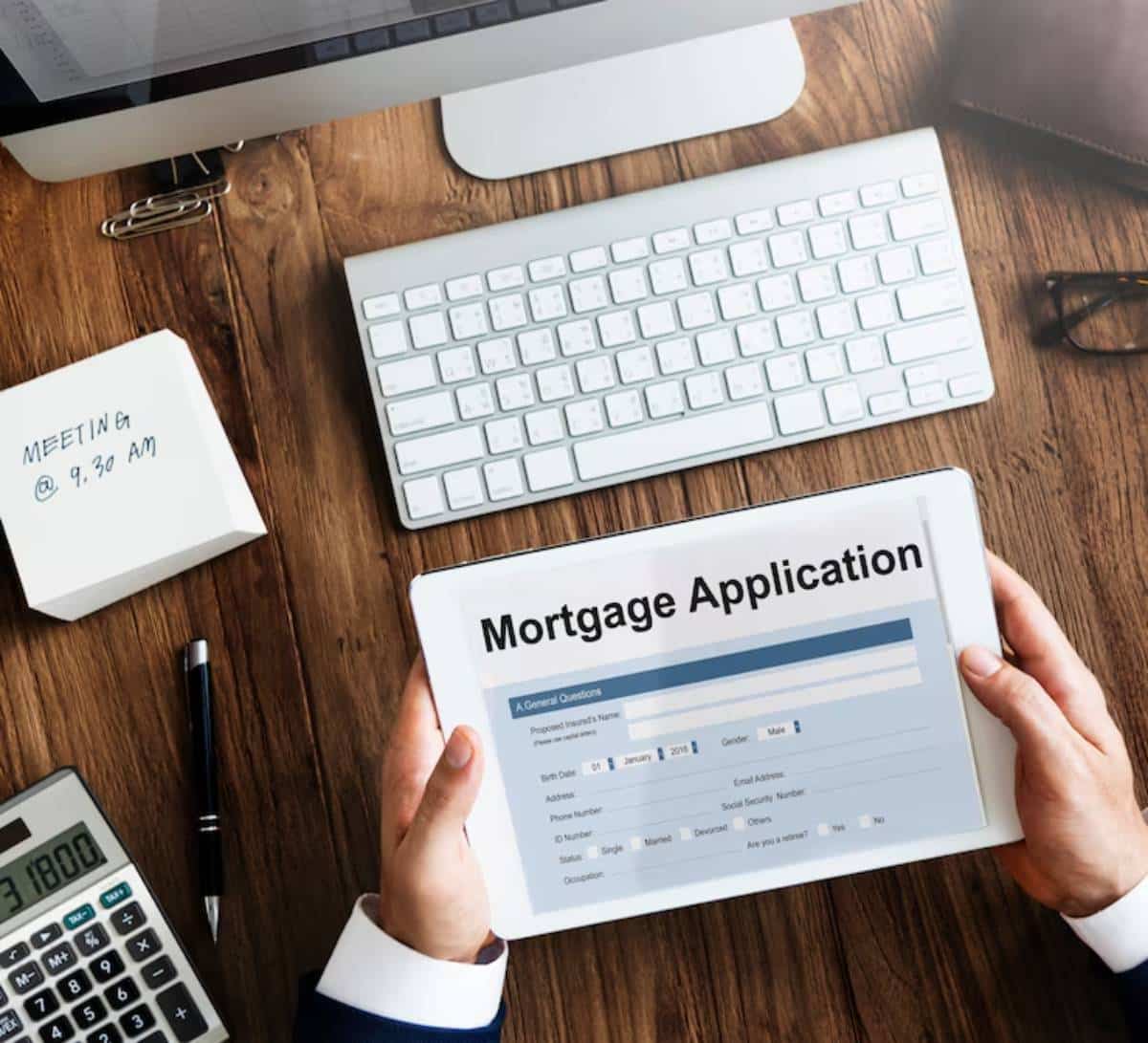 A person in a suit holds a tablet displaying a mortgage application form, with a computer and notepad on a wooden desk.